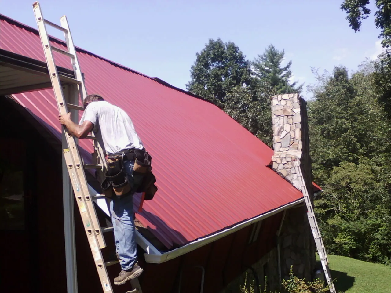 Roofer on a ladder beside a red metal roof and stone chimney — Soffit & Fascia Repair by Roofing Brothers in Spring Creek